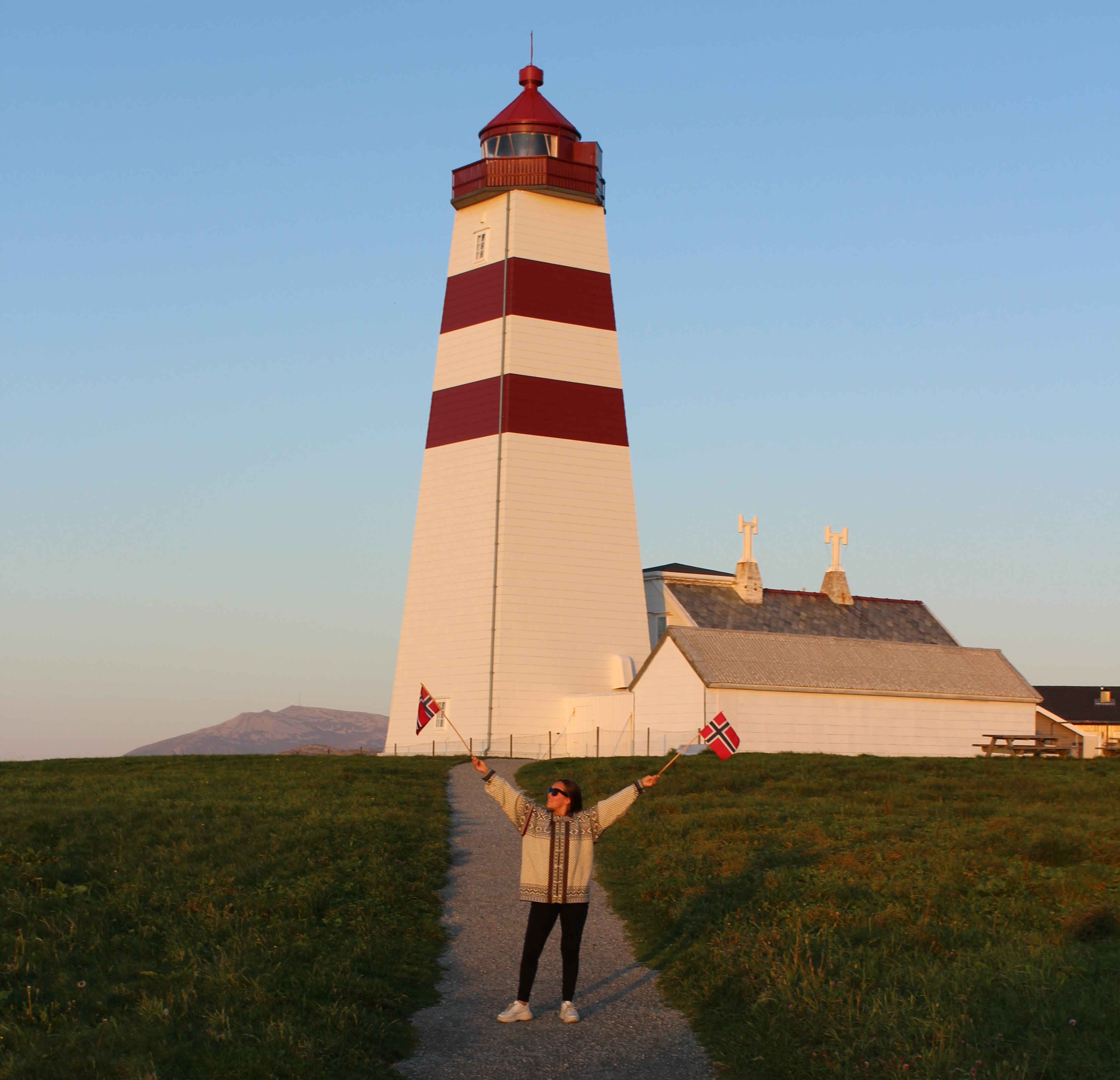 Image showing the Alnes Lighthouse in the background and a woman standing in front holding two Norwegian flags, one in each hand.